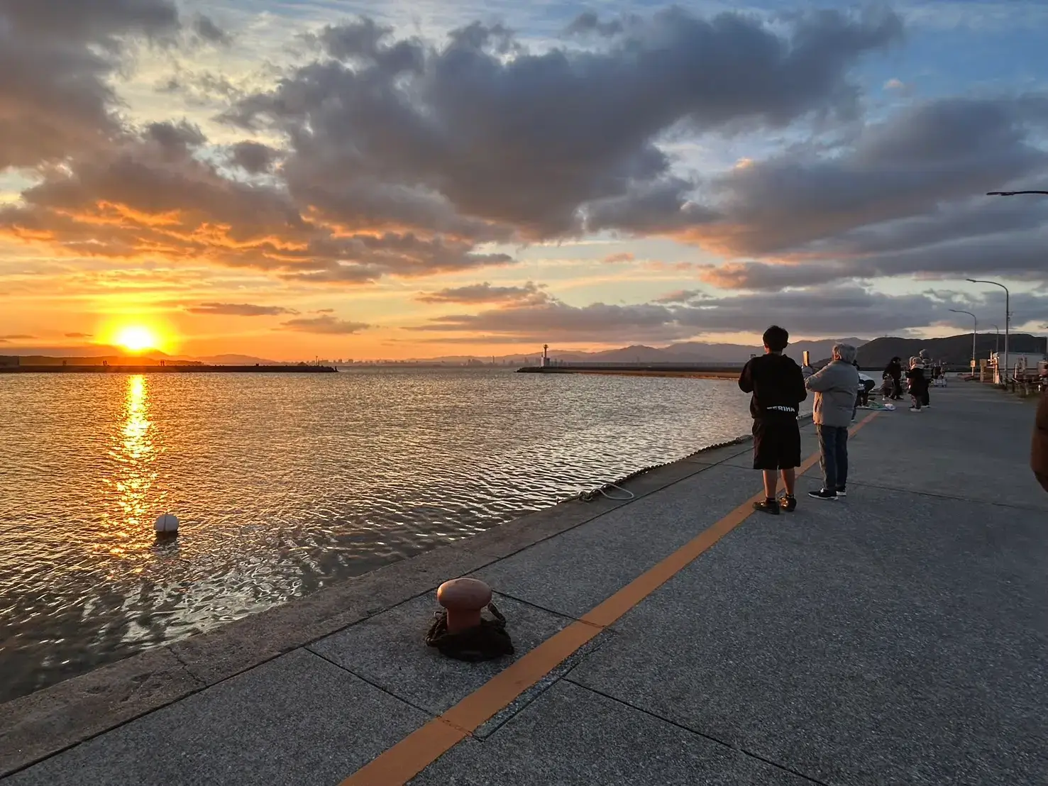 志賀島の岸壁で初日の出を眺める人々の後ろ姿。遠くにはアイランドシティのビル群と福岡市街地のスカイラインが広がる。